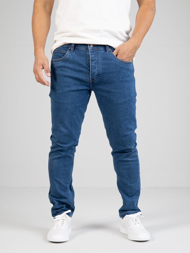 A person wearing a white t-shirt, Ganbaru Stretch Jeans by ID Menswear in a slim-tapered fit, and white sneakers stands with one hand in their pocket against a plain background. The jeans have a classic five-pocket design.
