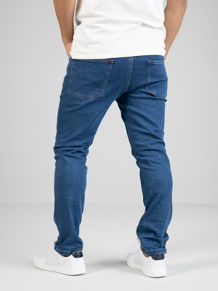 A person stands with their back to the camera, wearing ID Menswears Ganbaru Stretch Jean in a slim-tapered fit and a white T-shirt. They complete the look with white sneakers featuring dark accents, all set in a well-lit, plain environment.