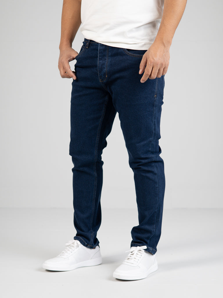 Wearing a white T-shirt and white sneakers, a person poses against a plain white background. They highlight the dark blue Ganbaru Stretch Jean by ID Menswear, featuring a slim-tapered fit that flatters any silhouette.