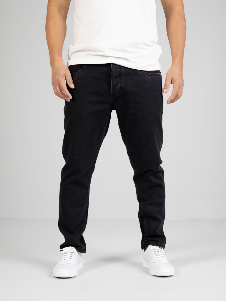 A person in a white t-shirt, ID Menswears black Ganbaru Stretch Jean, and white sneakers stands against a light gray background. The photo showcases their casual yet effortlessly stylish look from shoulders to feet.