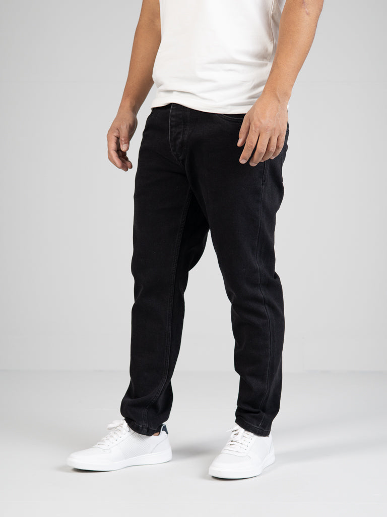 A person in a white T-shirt and ID Menswears Ganbaru Stretch Jeans stands casually against a plain gray background. With stylish white sneakers, the photo is cropped just below the neck, emphasizing their modern outfit.