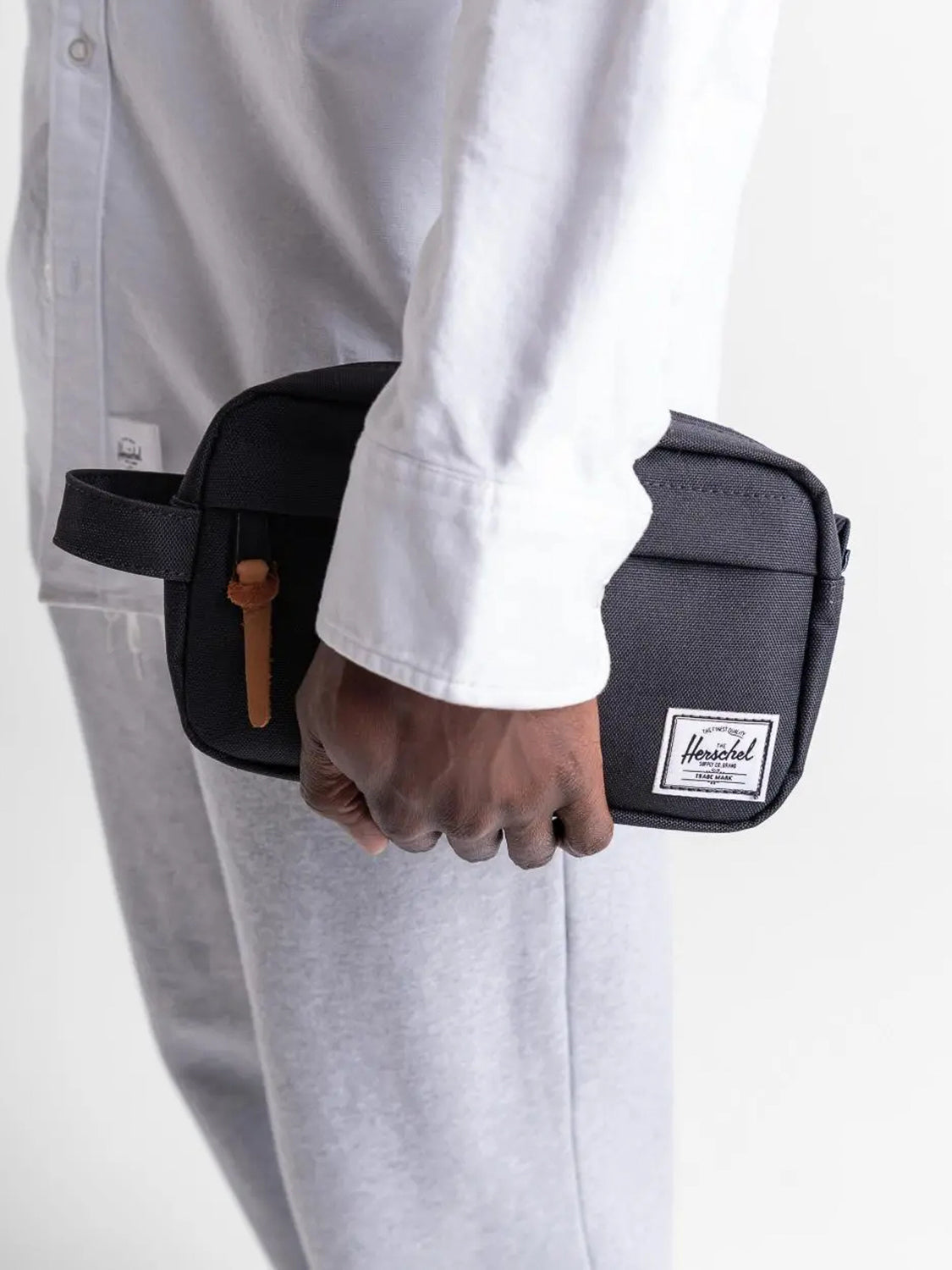 A person in a white shirt and light gray pants holds the Herschel Chapter Carry-On Travel Kit, featuring a leather zipper pull and white logo patch—ideal for stylish travel.