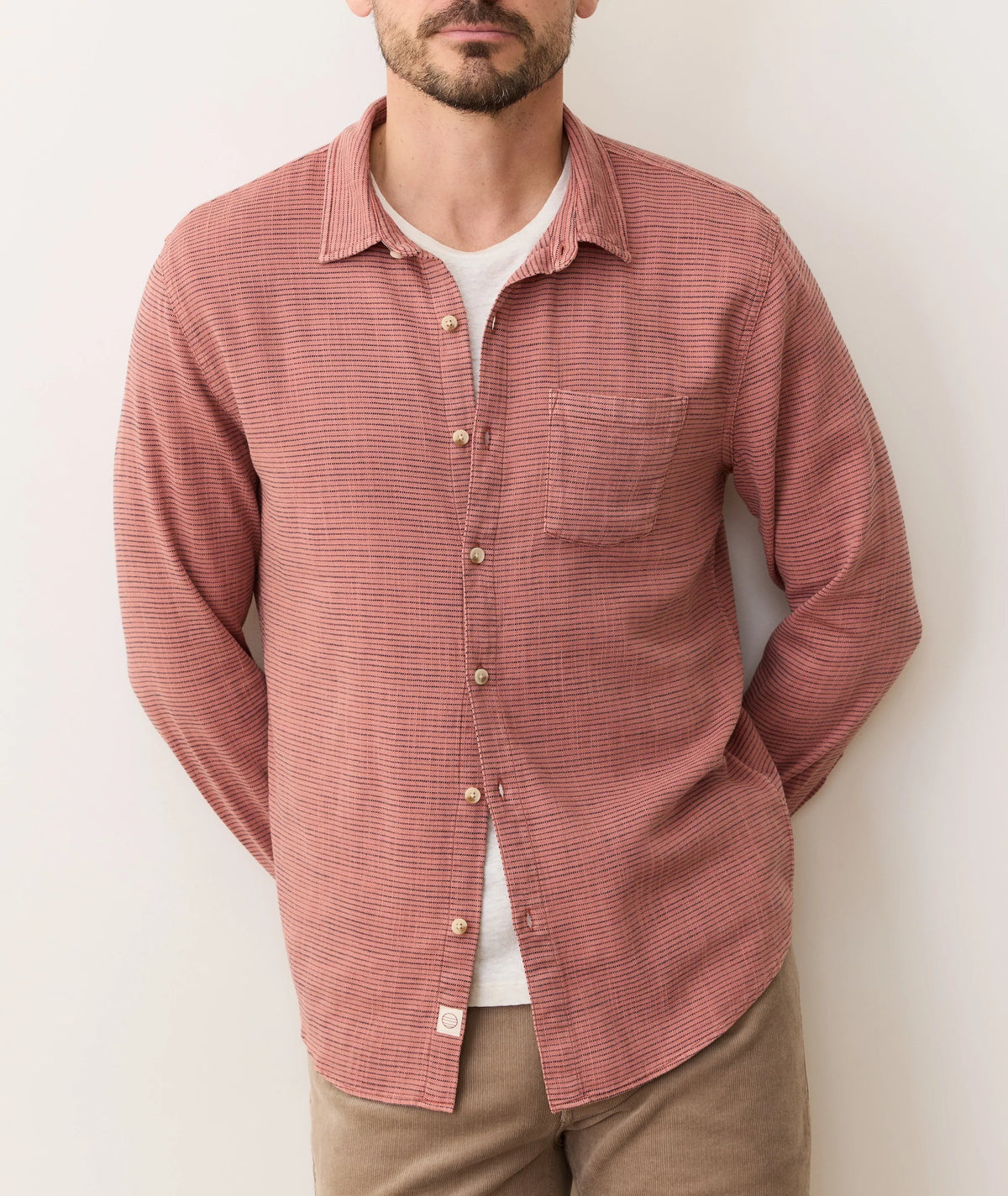A man stands against a plain light wall wearing a Marine Layer Stretch Selvage Long Sleeve Shirt in red and white check over a white tee with tan pants. His hands are behind his back, and his face is partially visible.