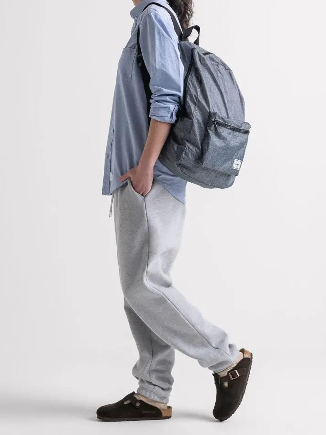 A person in a light blue button-up, gray sweatpants, and dark sandals stands in profile with one hand in their pocket, wearing a Herschel Packable Daypack with adjustable shoulder straps on their back.
