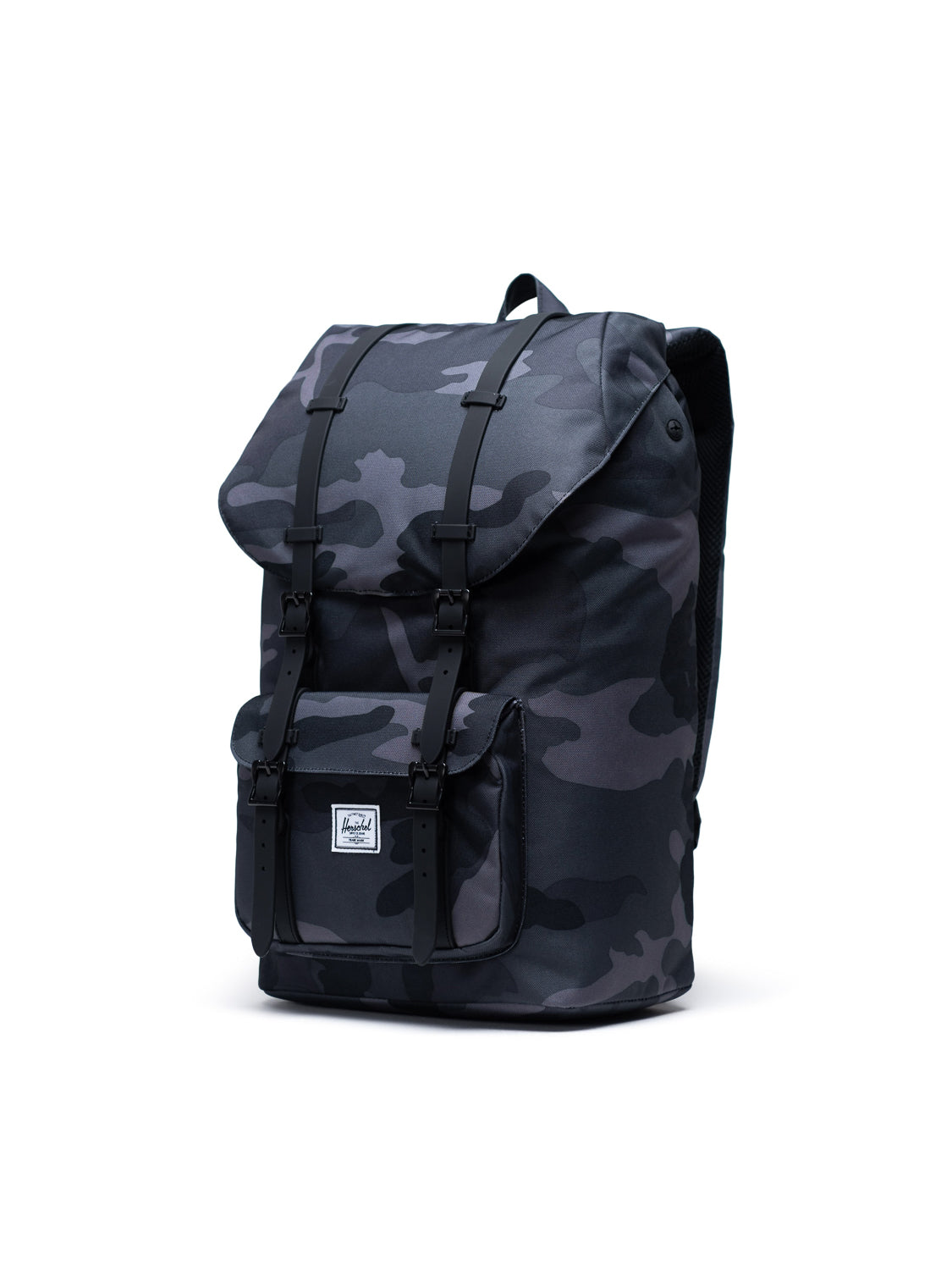 The Herschel Little America Iconic Backpack features a dark camo pattern, flap cover, black buckle straps, front pocket, and a white rectangular logo patch near the bottom. This backpack stands upright on a white background.