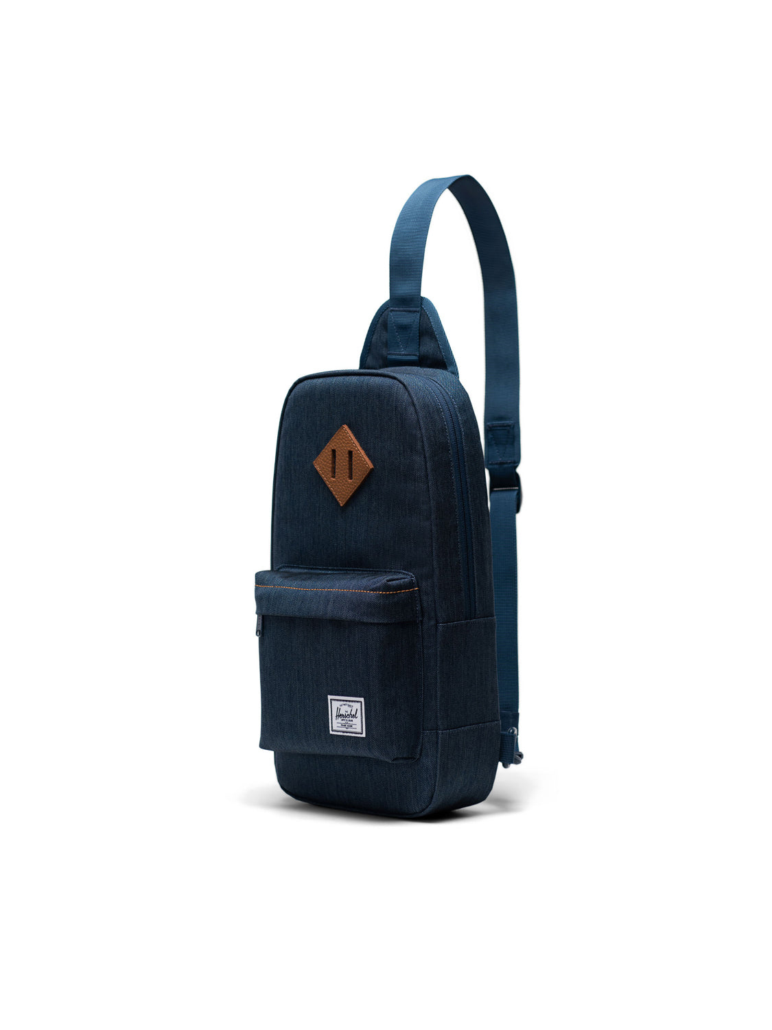 The Herschel Heritage Shoulder Bag is a blue, single-strap crossbody backpack with a front zip pocket, small rectangular bottom label, and a brown diamond-shaped patch on the upper front, shown on a white background.