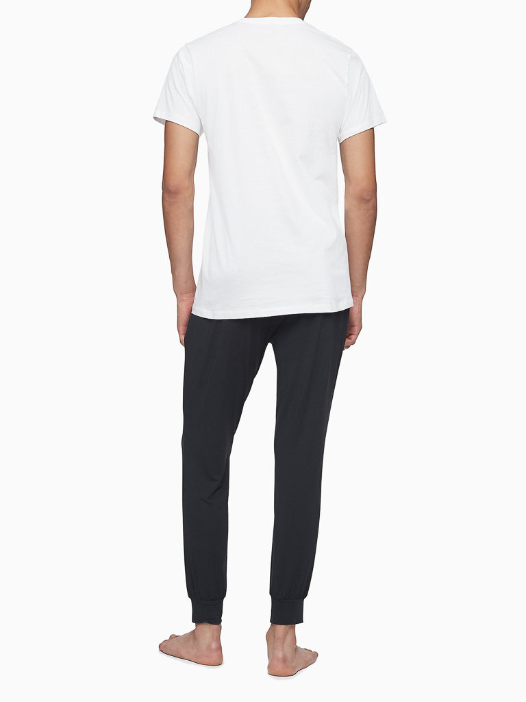 A person stands barefoot facing away from the camera, wearing a white Calvin Klein NB4051 3-Pack Cotton Classic Crewneck Tee and black jogger-style pants by Calvin Klein against a white background.