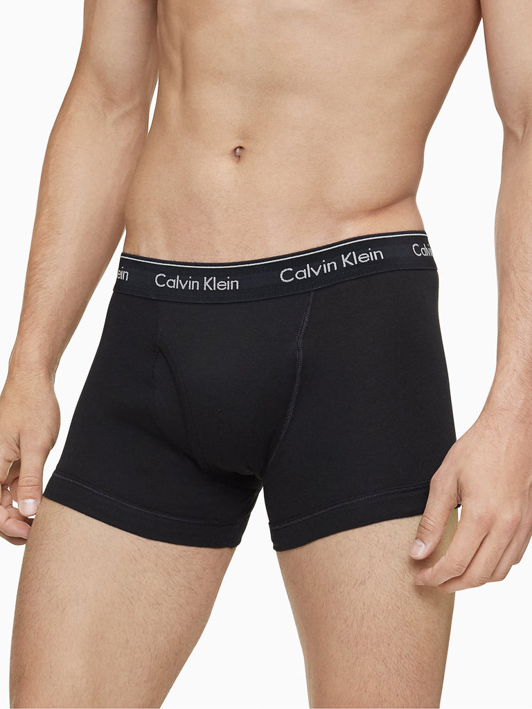 A person models Calvin Klein's NB4002 3-Pack Cotton Classic Fit Trunk in black, featuring the signature white Calvin Klein logo waistband. Photo shows mid-torso to mid-thigh, with arms at their sides.
