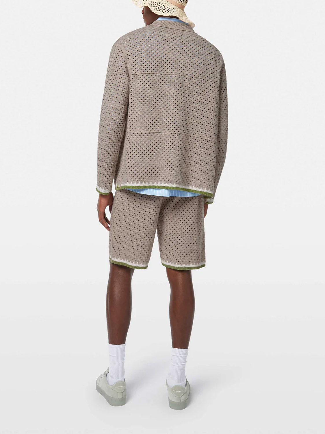 A person stands with their back to the camera, wearing SCOTCH AND SODA’s Crochet Long Sleeve Shirt 180084 SU25 and matching shorts, white socks, light gray shoes, and a beige hat. The plain white background creates an artistic look.