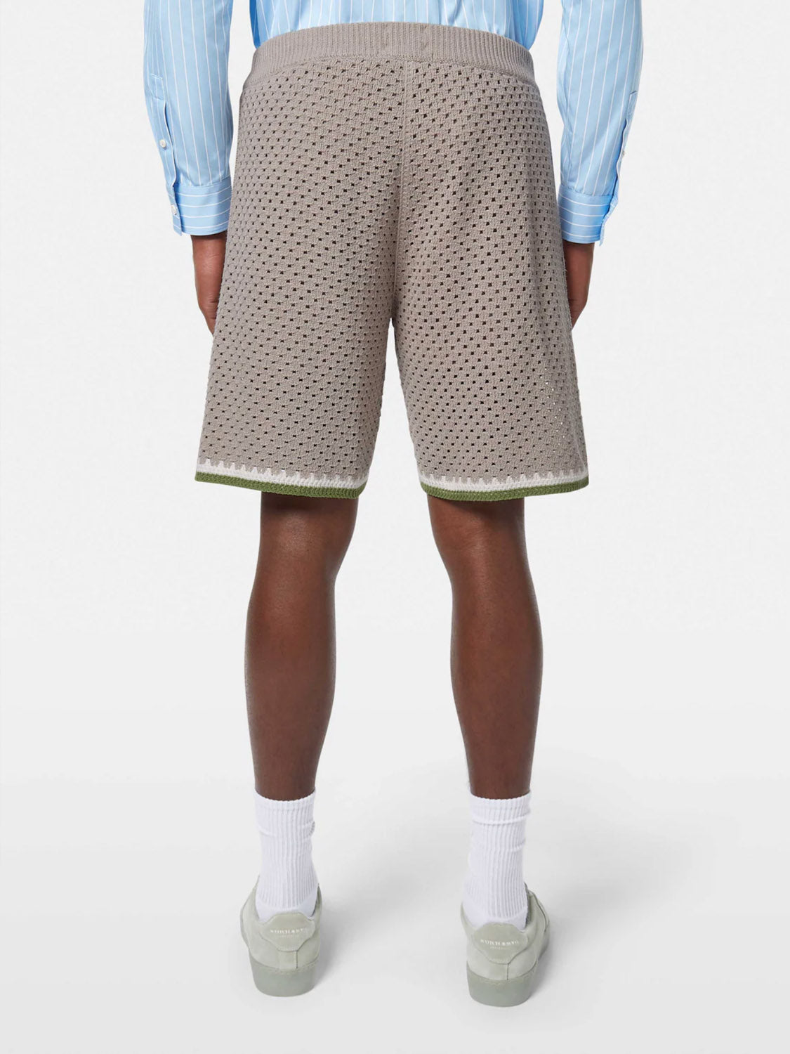 A person faces away, wearing SCOTCH AND SODA Crochet Knit Short 180344 SU25 in taupe with green trim, paired with a blue and white striped shirt, white socks, and light shoes against a plain white background.