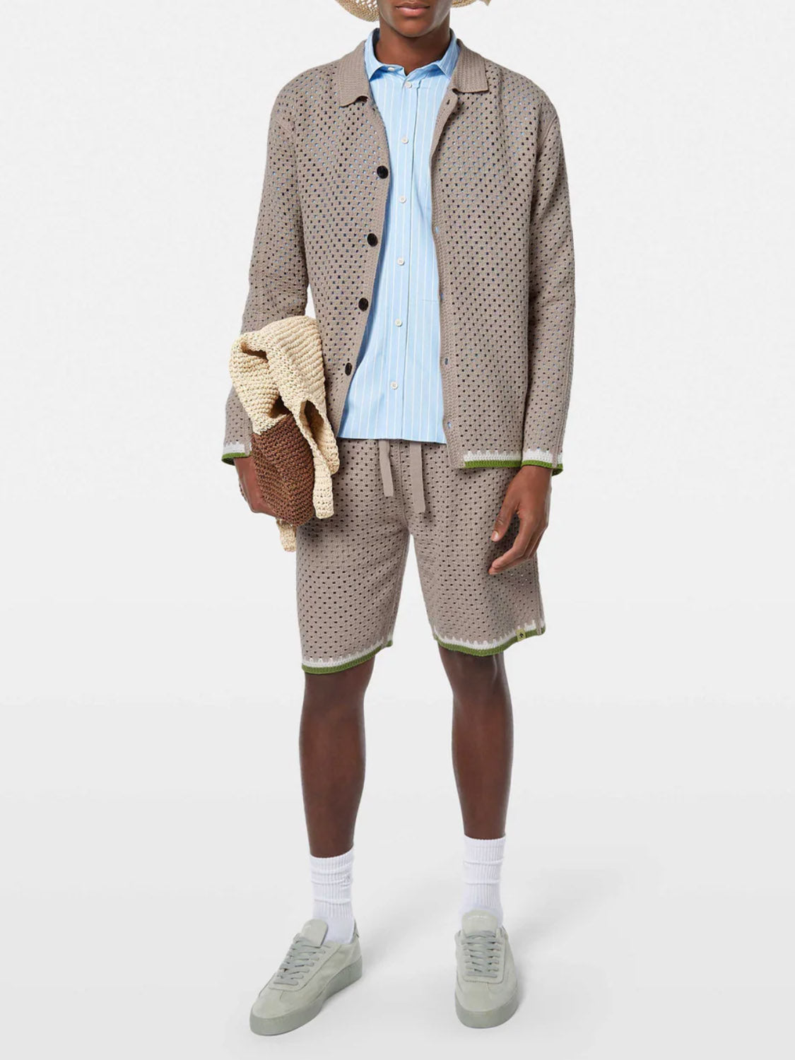 A person stands against a plain light background, wearing SCOTCH AND SODA Crochet Knit Short 180344 SU25 paired with a tan polka-dotted jacket, blue and white striped shirt, white socks, gray sneakers, and holding knitted items. Head is out of frame.