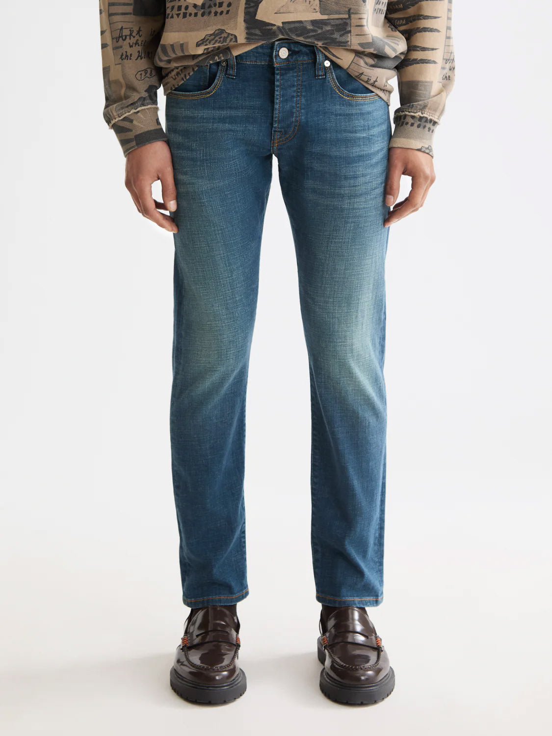 A person wearing Scotch & Soda 180872 Ralston Regular Slim Denim jeans, a patterned long-sleeve shirt, and dark brown dress shoes stands against a plain white background.