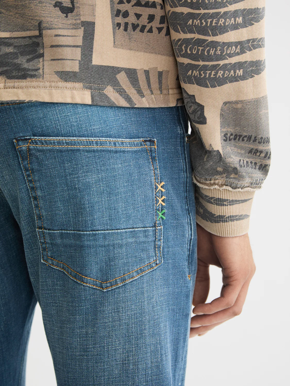 A close-up shows a person wearing Scotch & Soda's 180872 Ralston Regular Slim Denim jeans with colored cross-stitch near the back pocket and a beige patterned top featuring text like AMSTERDAM. Only part of their torso and hand are visible.