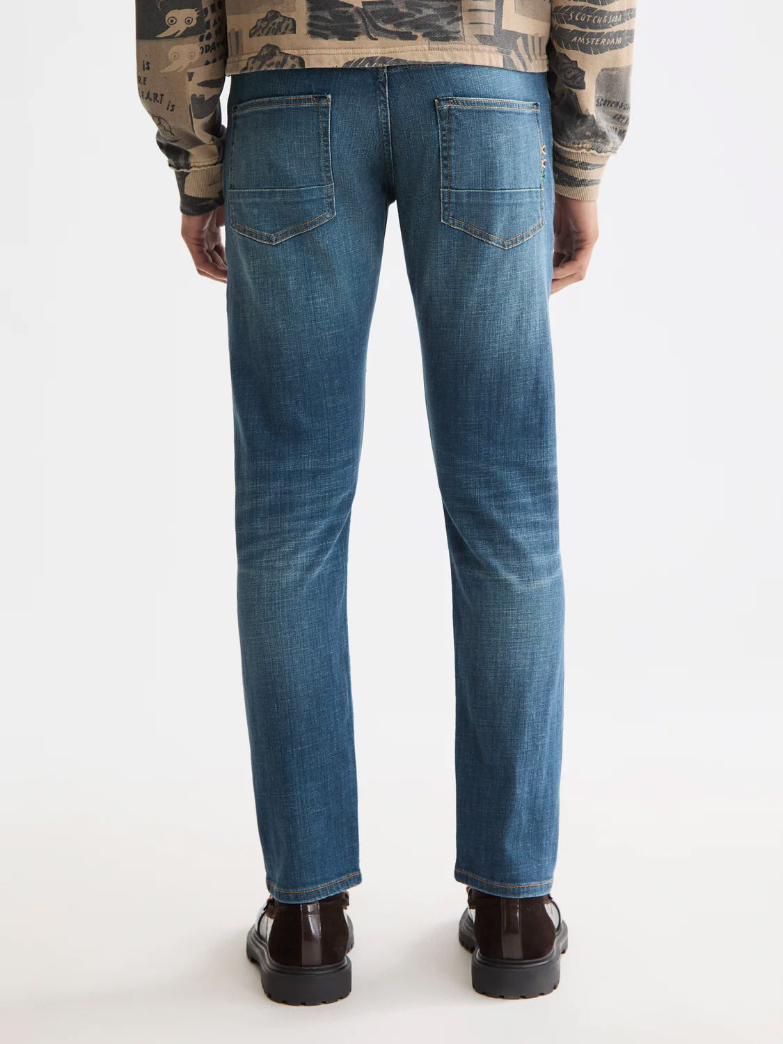 A person stands with their back to the camera, wearing Scotch & Soda 180872 Ralston Regular Slim Denim jeans in blue and brown shoes, paired with a patterned long-sleeve top. The jeans have back pockets and a slightly faded finish. Background is plain white.