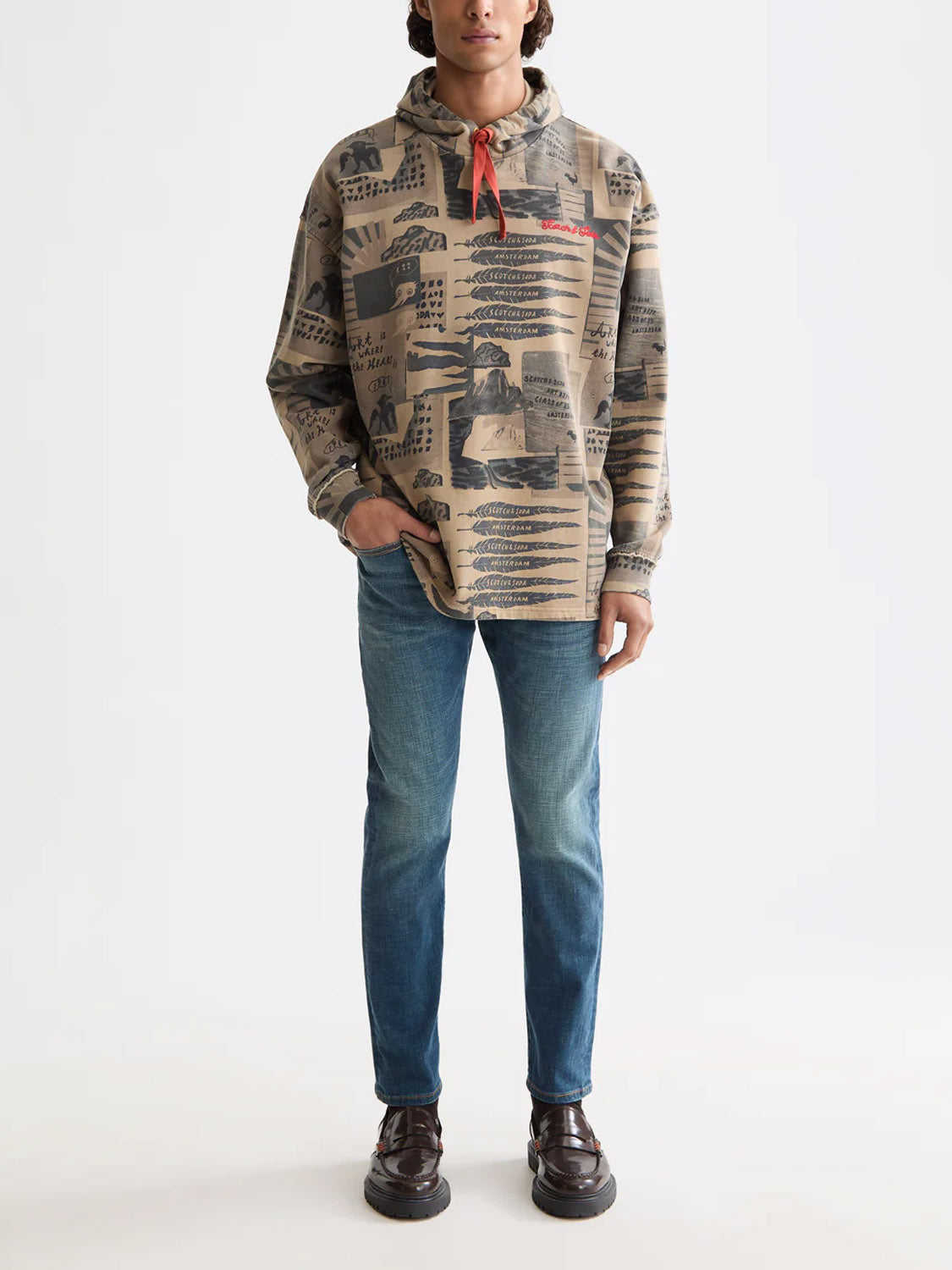 A person stands facing forward against a plain white background, wearing a tan hoodie with black graphics, dark brown shoes, and Scotch & Soda 180872 Ralston Regular Slim Denim jeans in blue. Their left hand is in their pocket.