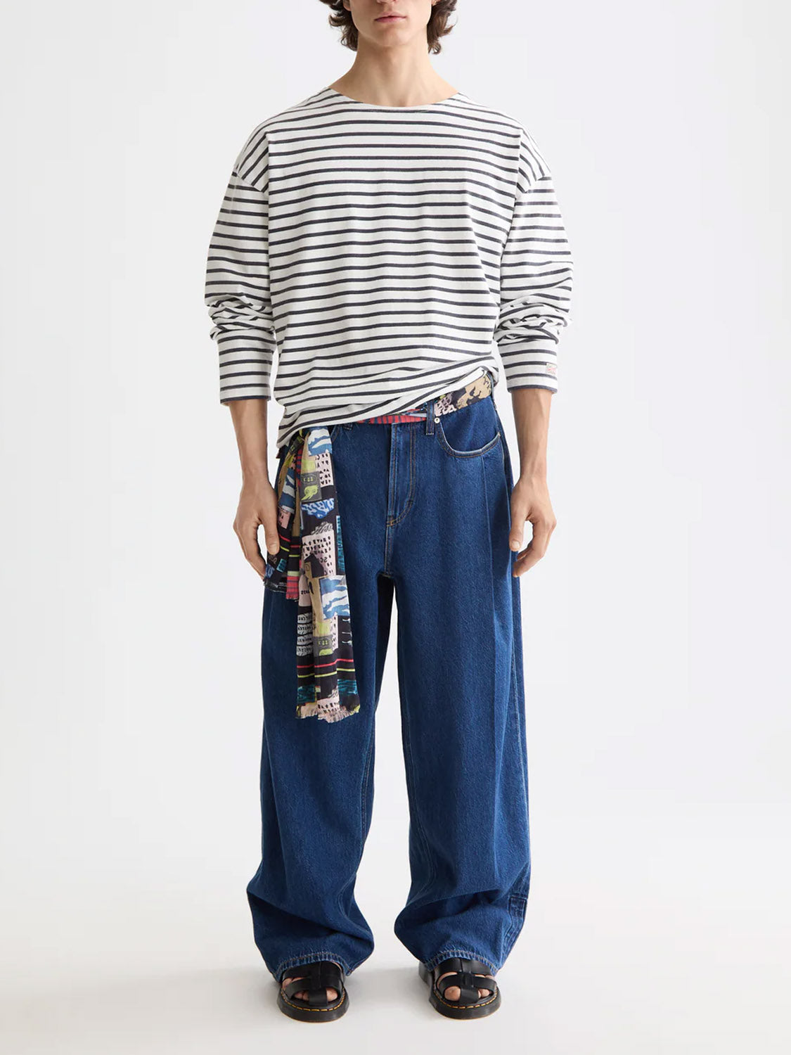 A person in a Scotch & Soda 180878 Denim Washed Long Sleeve Breton Tee with black-and-white stripes, wide-leg jeans, black sandals, and a colorful patterned scarf tied at the waist stands against a plain white background.