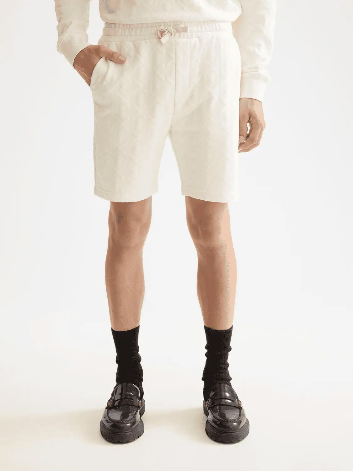 A person wearing Scotch & Soda's 181210 Jacquard Terry Sweatshorts and a matching white sweatshirt, paired with black socks and loafers, stands hands in pockets against a plain white background.
