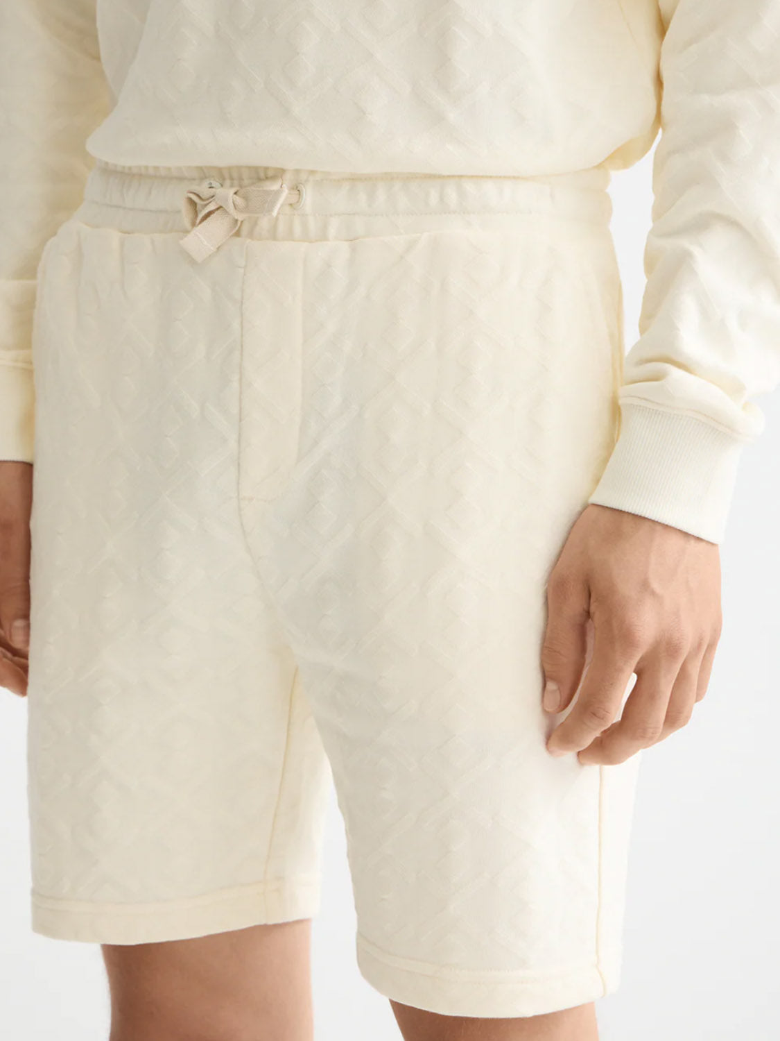 A person wears Scotch & Soda 181210 Jacquard Terry Sweatshorts in cream, featuring a relaxed fit, drawstring waist, and subtle diamond pattern. Only the lower torso and upper legs are visible; paired with a matching sweatshirt.