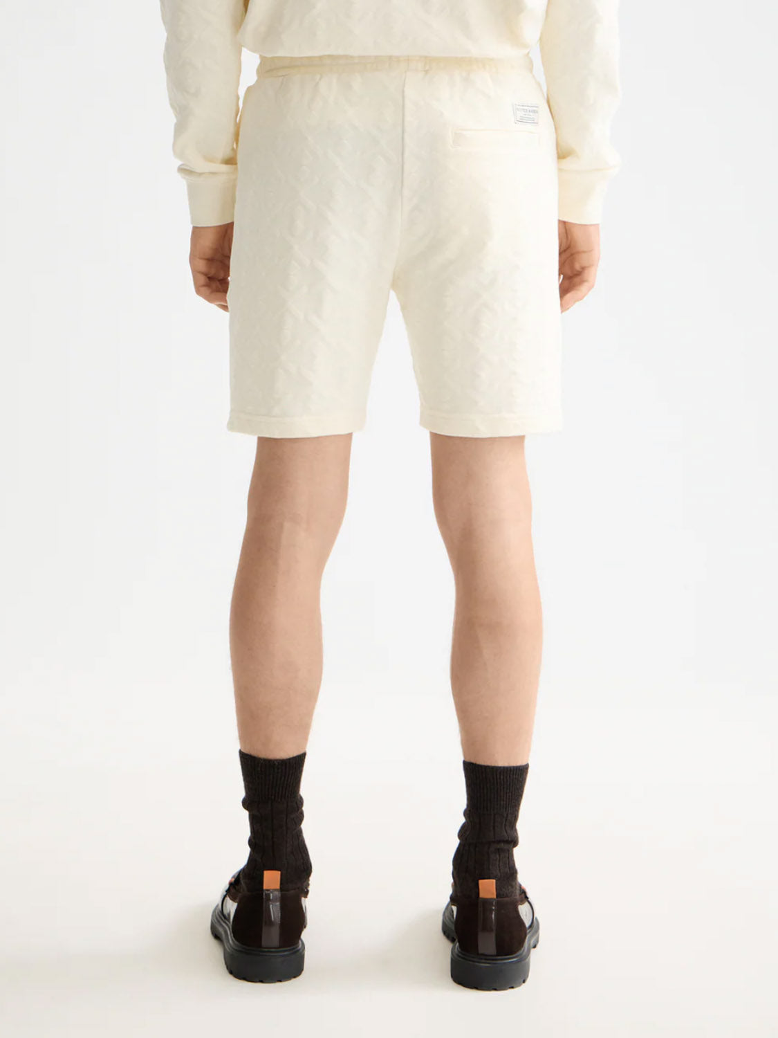 A person faces away from the camera, wearing Scotch & Soda 181210 Jacquard Terry Sweatshorts with a matching sweatshirt, dark socks, and black sandals with brown accents, set against a plain white background.