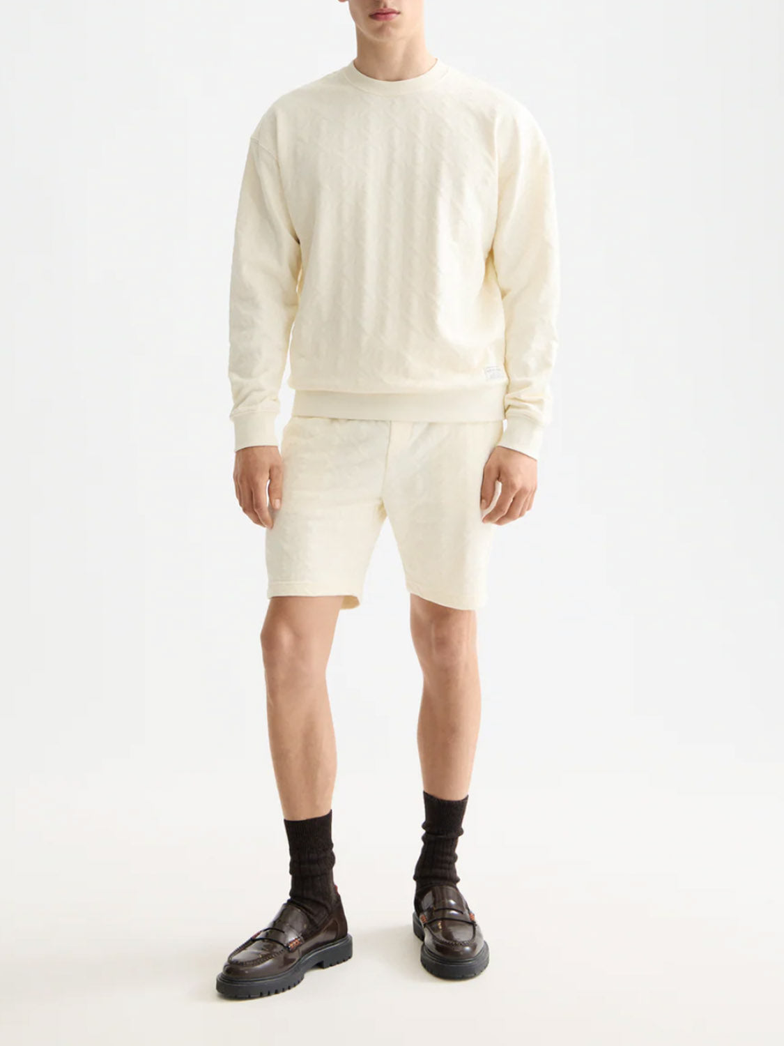 A person models the Scotch & Soda 181210 Jacquard Terry Sweatshorts with a coordinating sweatshirt, black socks, and dark brown loafers. They face forward against a plain white background, head partially out of frame.