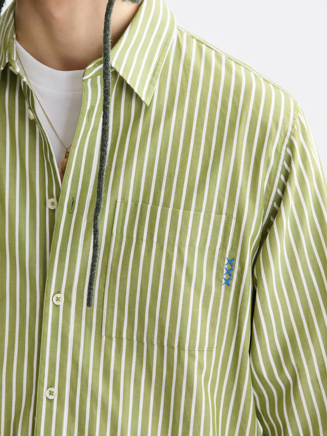 A close-up of a person wearing Scotch & Soda’s 181232 Striped Poplin Shirt in green and white, made from 100% cotton, featuring a front pocket with blue embroidery, over a white undershirt. Part of a necklace and dark drawstring are visible.