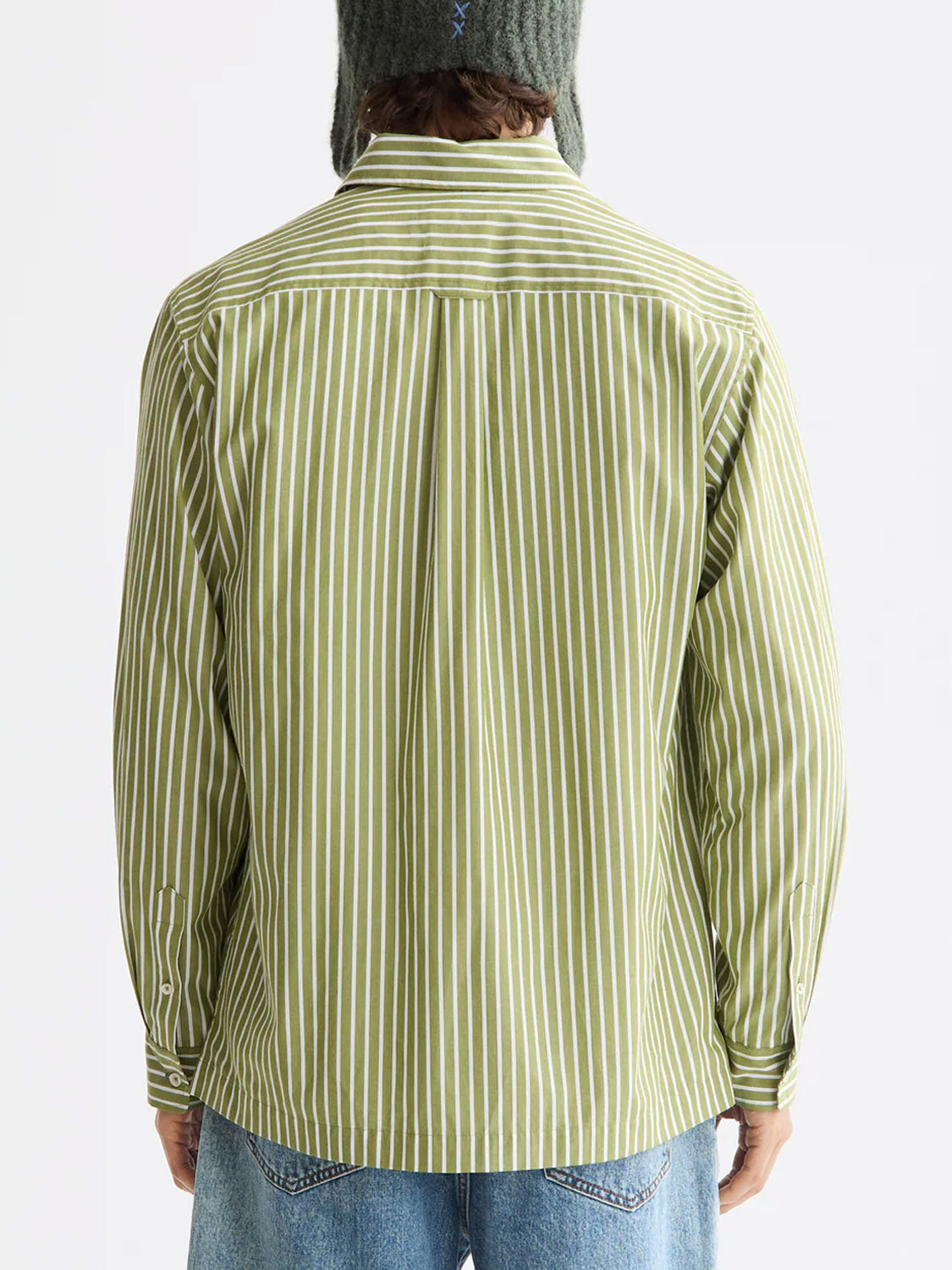 A person is shown from the back wearing the Scotch & Soda 181232 Striped Poplin Shirt in green and white vertical stripes, 100% cotton, with long sleeves rolled down, blue jeans, and a knit hat.