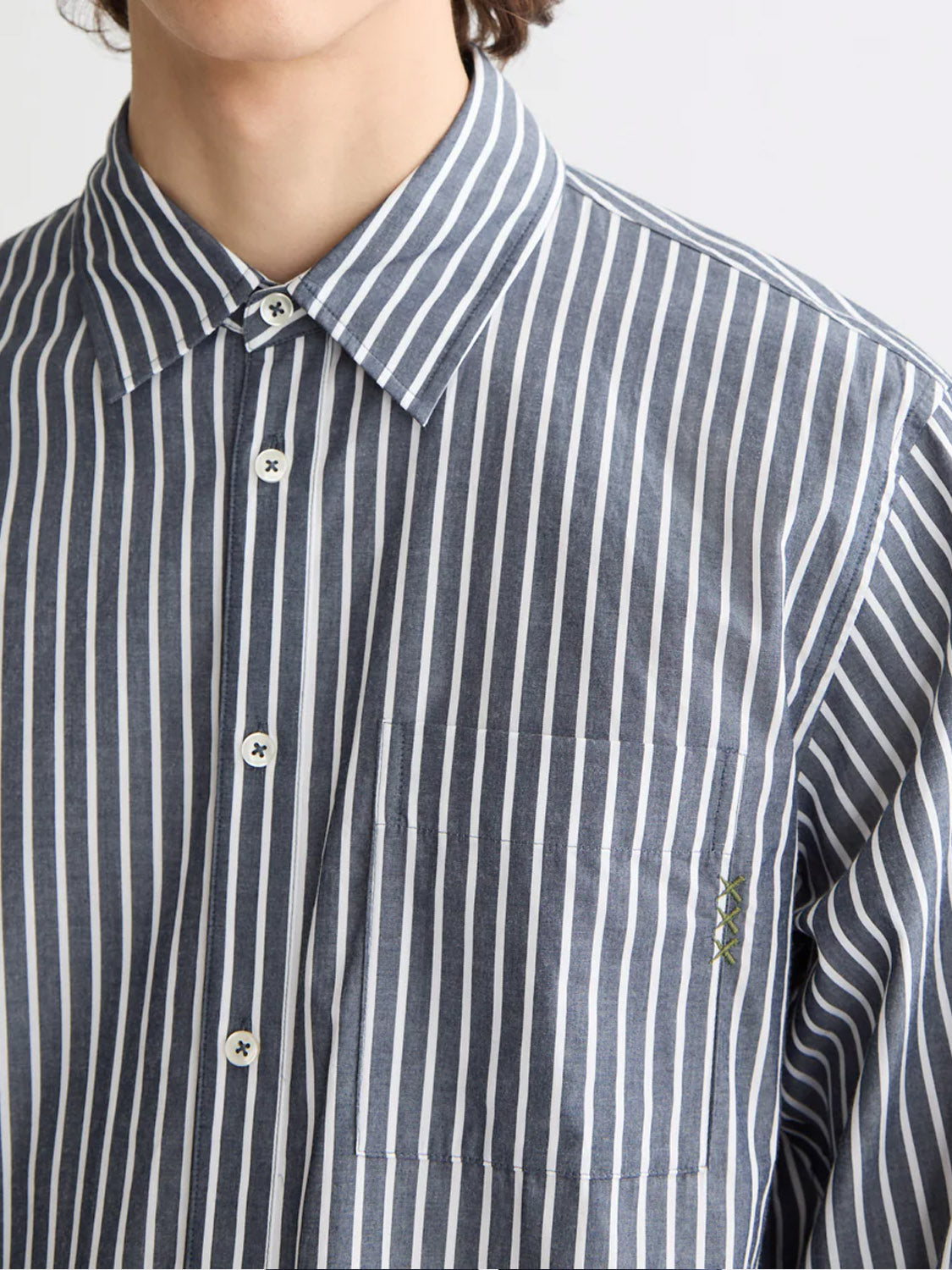 A person wears the Scotch & Soda 181232 Striped Poplin Shirt, a relaxed-fit, blue and white vertical stripe shirt in 100% cotton with white buttons and a chest pocket featuring a small green embroidered detail; upper torso partly visible.