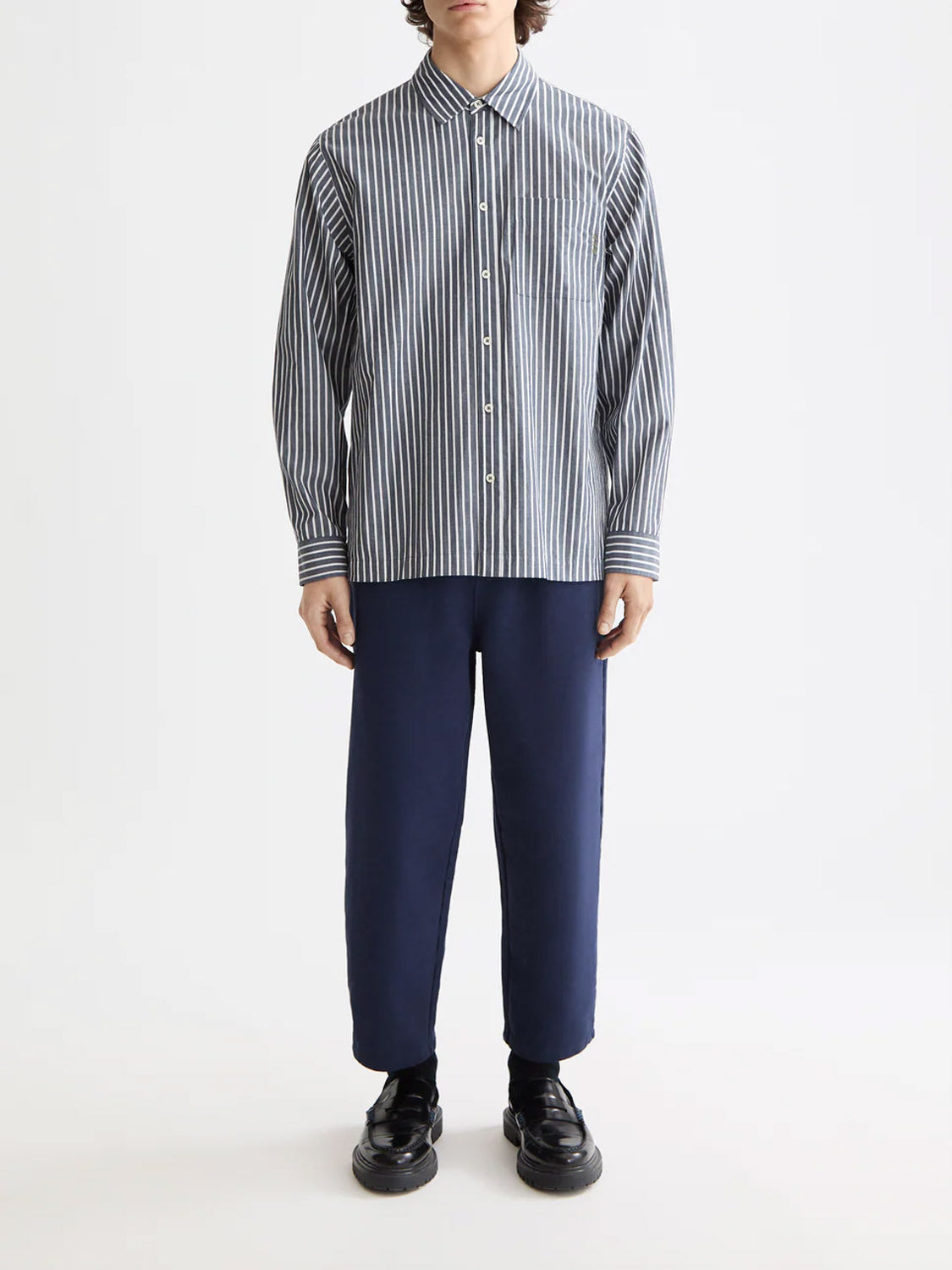 A person stands against a plain background wearing the Scotch & Soda 181232 Striped Poplin Shirt in blue and white, made from 100% cotton, paired with navy pants and black shoes. The person’s face is not visible.