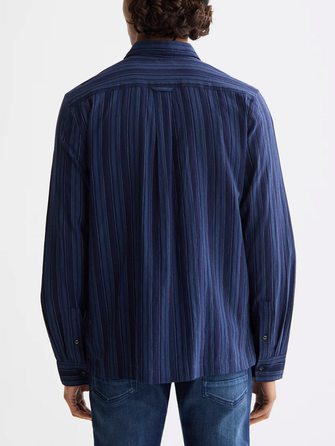 A person stands facing away, wearing the Scotch & Soda 181248 Textured Stripe Shirt—an indigo, 100% cotton shirt with vertical stripes—and blue jeans against a plain, light-colored background.