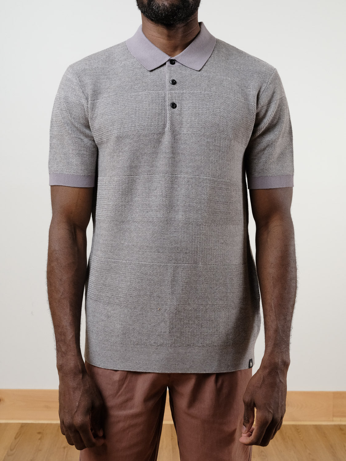 A man wears the SCOTCH AND SODA Knitted Tonal Striped Regular Polo, a classic fit, light gray 100% cotton shirt with collar and three buttons, paired with light brown pants against a plain light background.