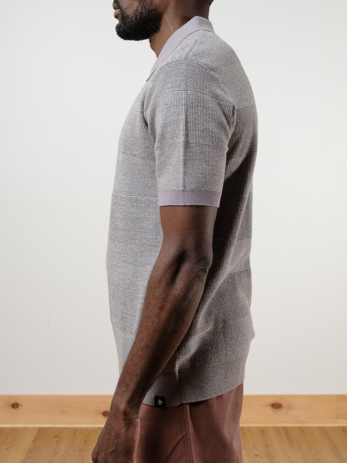 A man, partly out of frame, stands sideways on a wooden floor against a light background, wearing SCOTCH AND SODA's Knitted Tonal Striped Regular Polo in light gray and mauve pants.