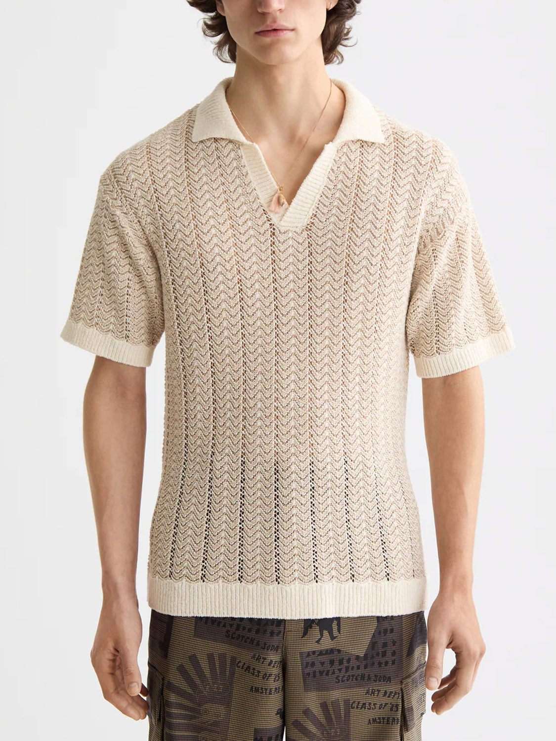 A person wears the Scotch & Soda 181322 Open Structured Knitted V-Neck Polo in cream, featuring a lightweight chevron pattern and ribbed collar, paired with brown printed shorts. The face is partially out of frame.