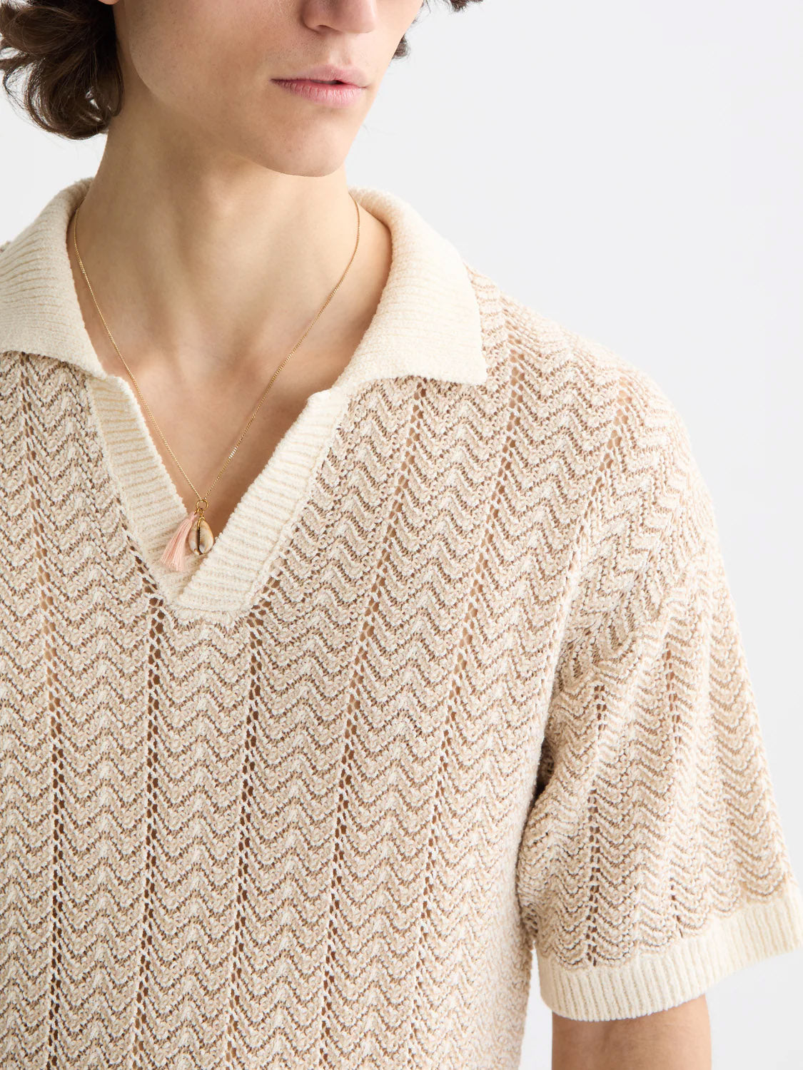 A person wears the Scotch & Soda 181322 - Open Structured Knitted V-Neck Polo in cream, featuring short sleeves and a wide collar, paired with a gold necklace, shown from shoulders up against a plain background.