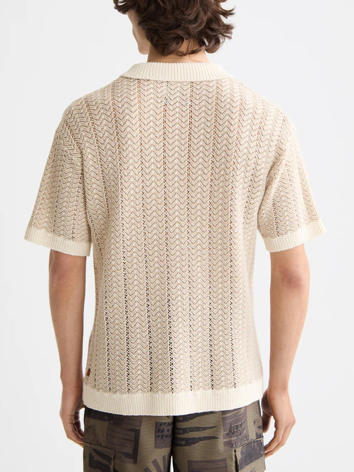 A person with wavy brown hair, seen from the back, wears the Scotch & Soda 181322 Open Structured Knitted V-Neck Polo in light beige with patterned camo shorts.