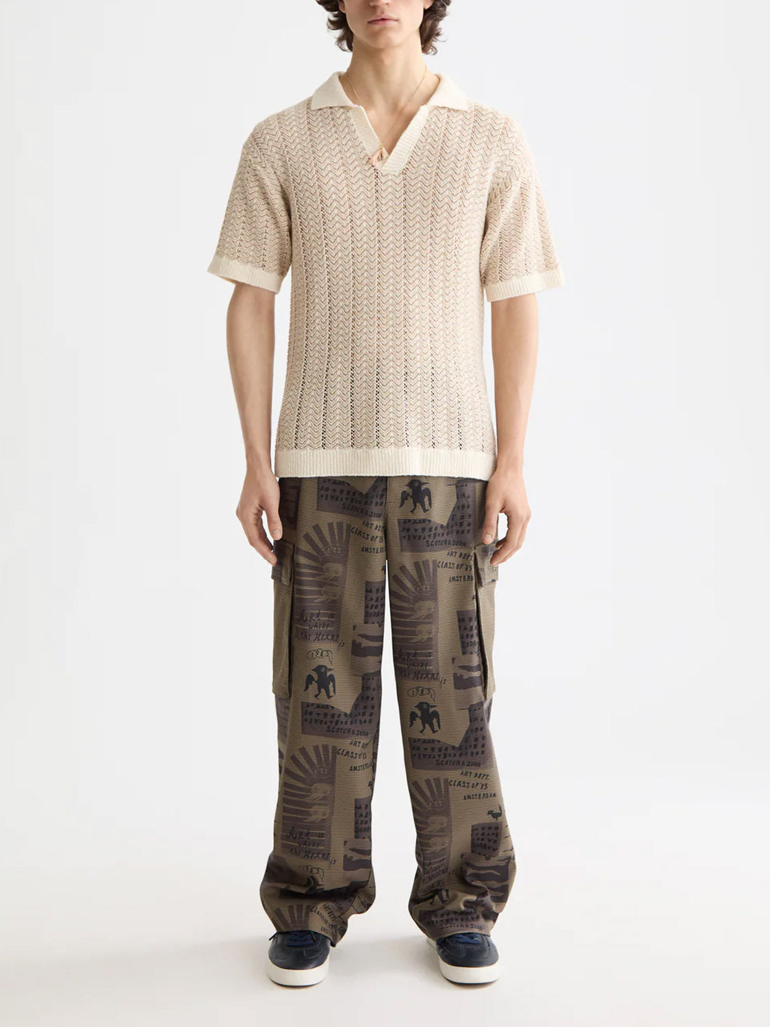 A person stands against a plain background wearing the Scotch & Soda 181322 Open Structured Knitted V-Neck Polo in cream, paired with loose olive cargo pants featuring bold black graphics and dark sneakers.