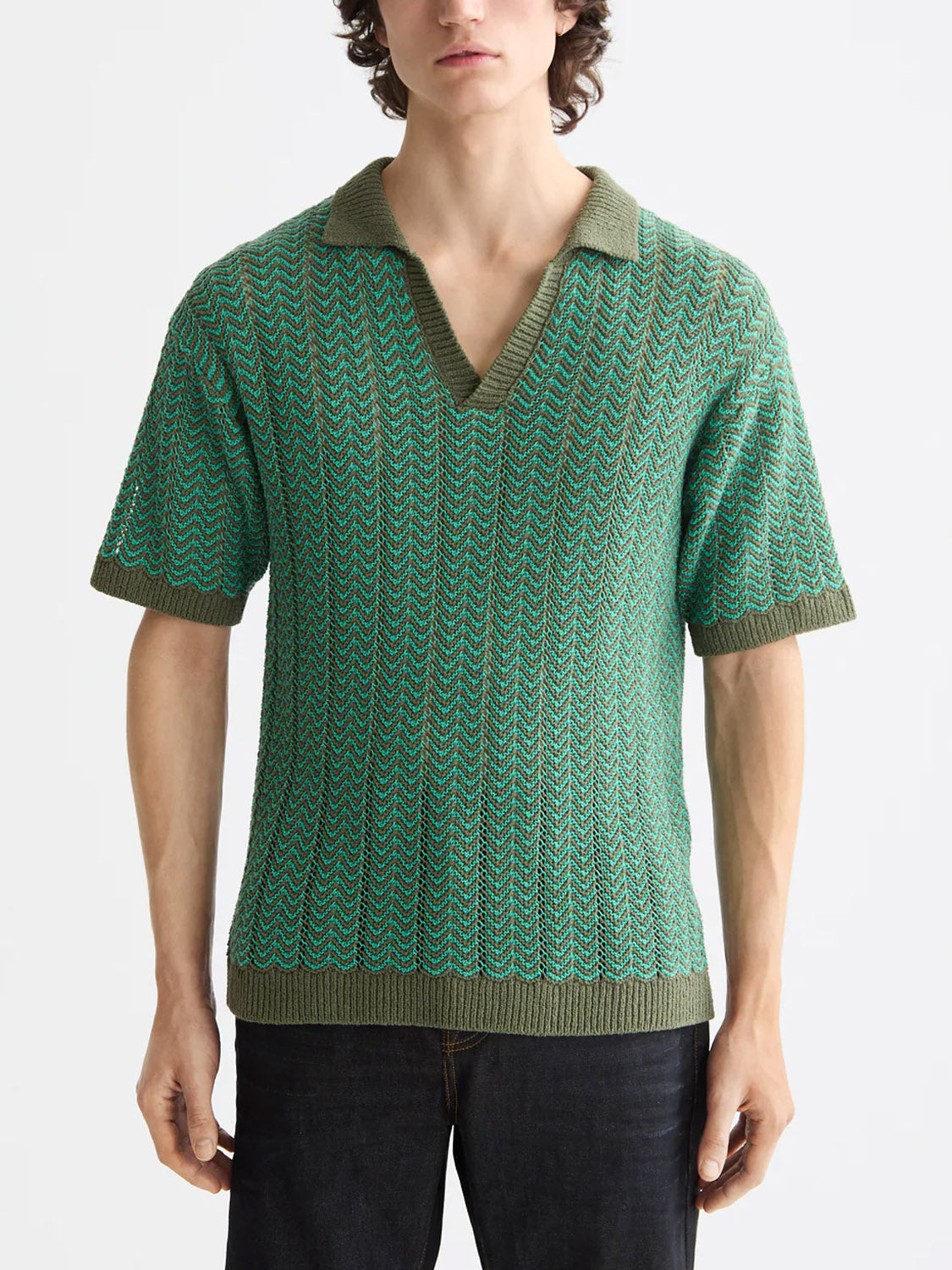 A person wears the Scotch & Soda 181322 Open Structured Knitted V-Neck Polo, a short-sleeved green shirt with collar, paired with dark pants, standing against a plain light background.