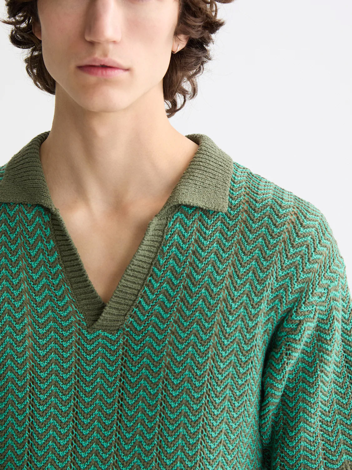 A person with wavy brown hair models the Scotch & Soda 181322 Open Structured Knitted V-Neck Polo, a lightweight green and teal zigzag-patterned sweater, shown from the upper torso against a plain light background.