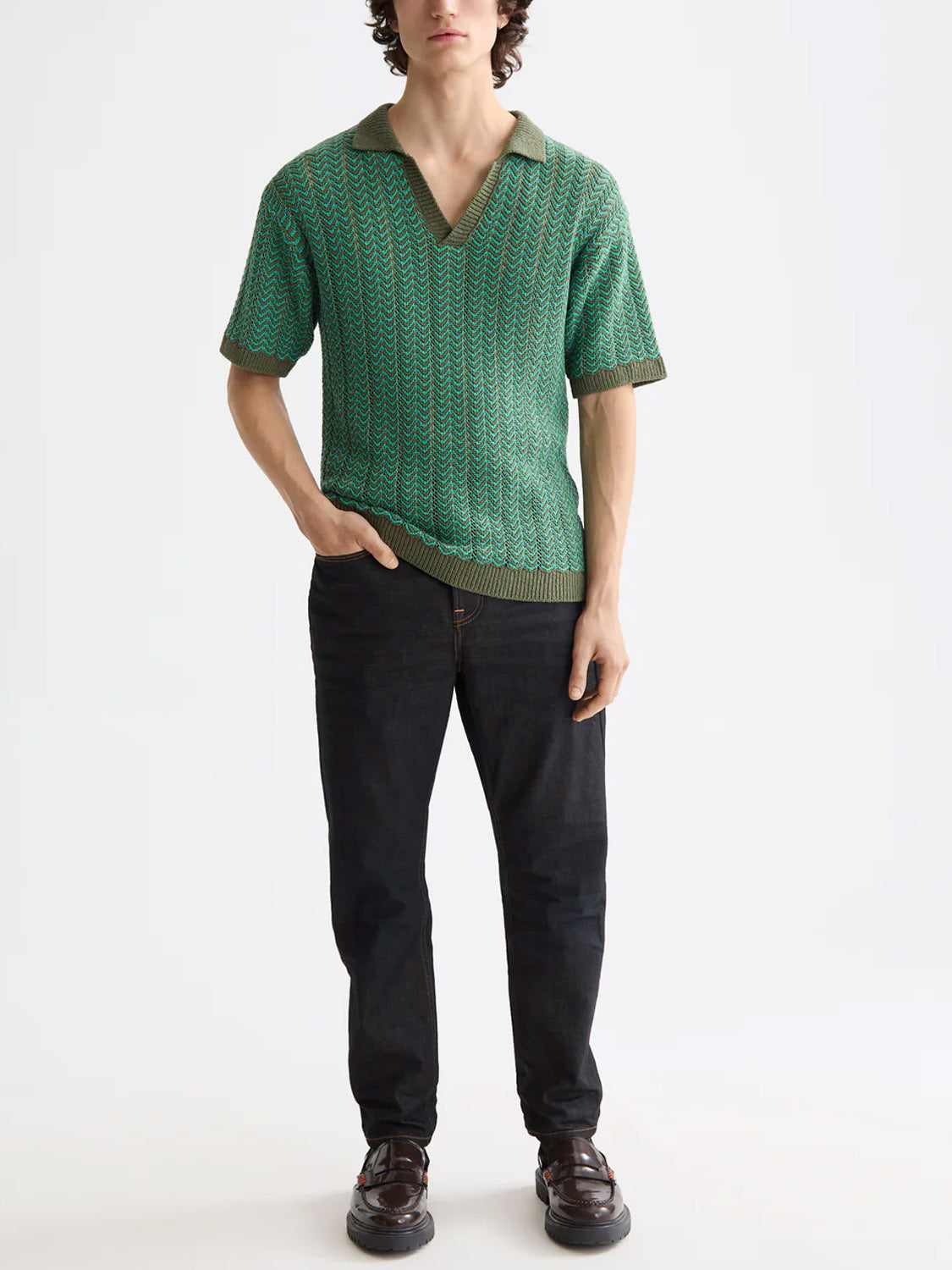A person stands against a plain background wearing the Scotch & Soda 181322 Open Structured Knitted V-Neck Polo in green, paired with dark jeans and dark brown shoes. One hand is in their pocket; their face is not fully visible.