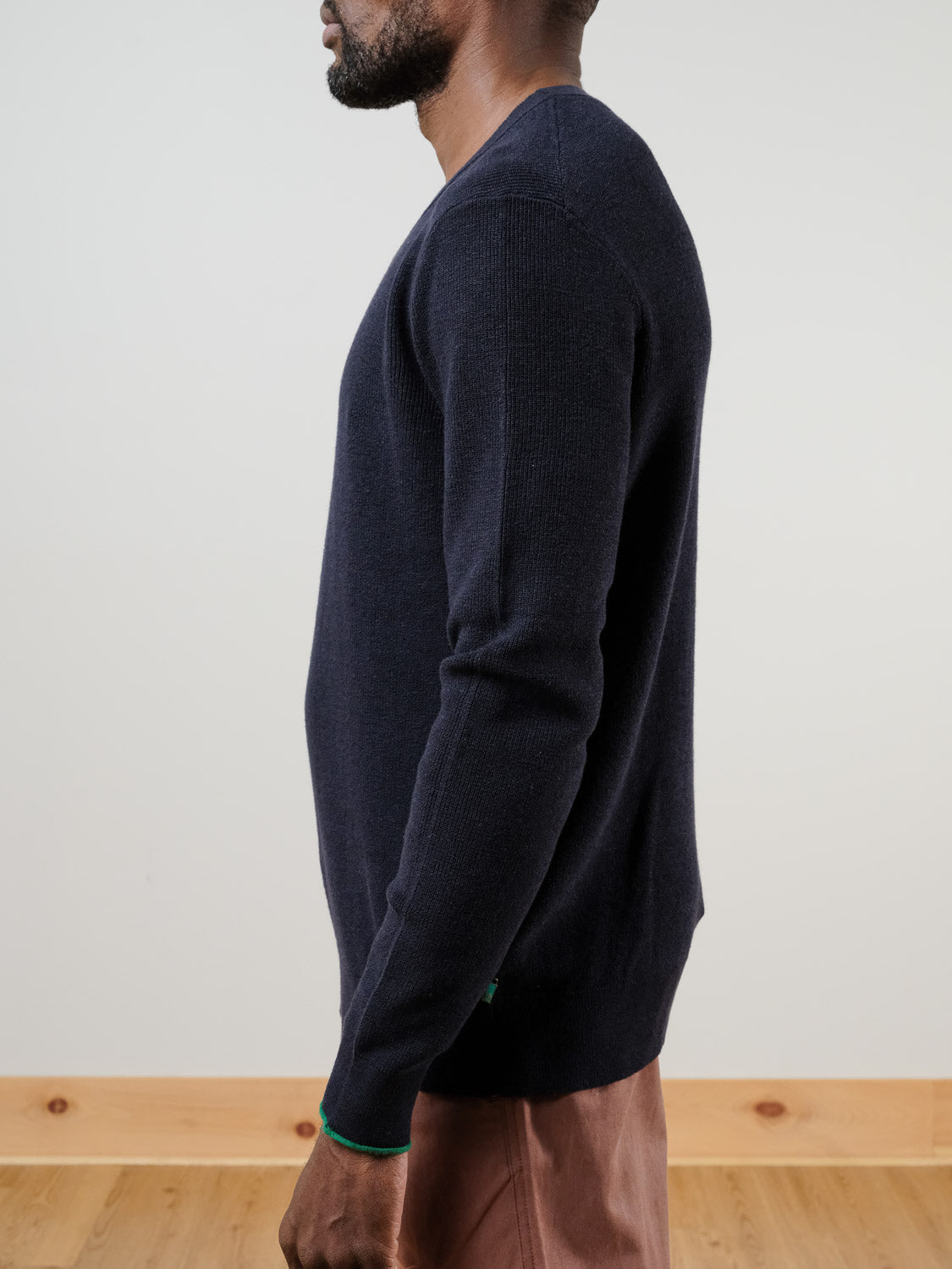 A man in profile stands on a wooden floor with minimal decor, wearing SCOTCH AND SODA’s Linen Blend Crew Neck in navy blue paired with light brown pants.