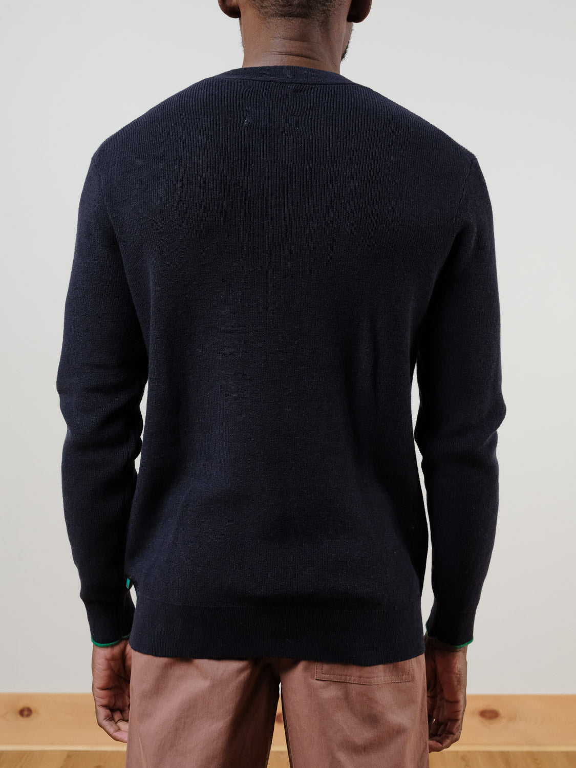 A person faces away from the camera, wearing a SCOTCH AND SODA Linen Blend Crew Neck in dark navy and light brown pants. The background features a plain light wall and wooden floor.