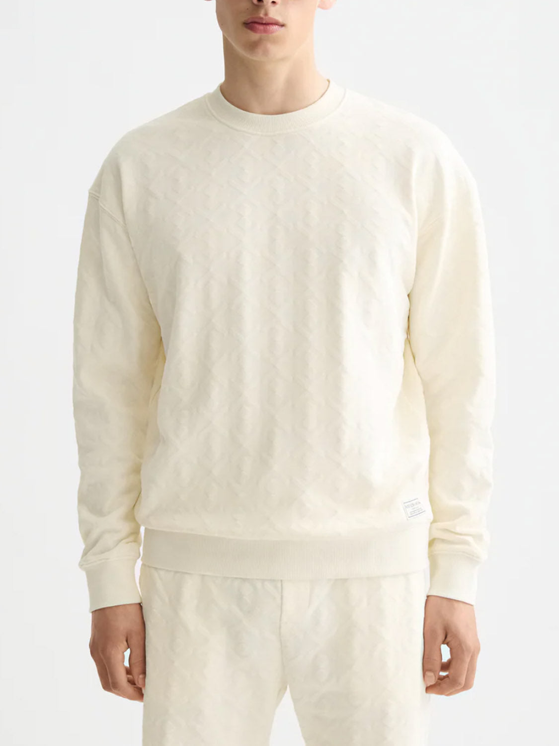 A person models the Scotch & Soda 181532 Jacquard Terry Sweatshirt in off-white, featuring long sleeves and a subtle textured pattern, paired with matching pants against a plain white background for an understated look.