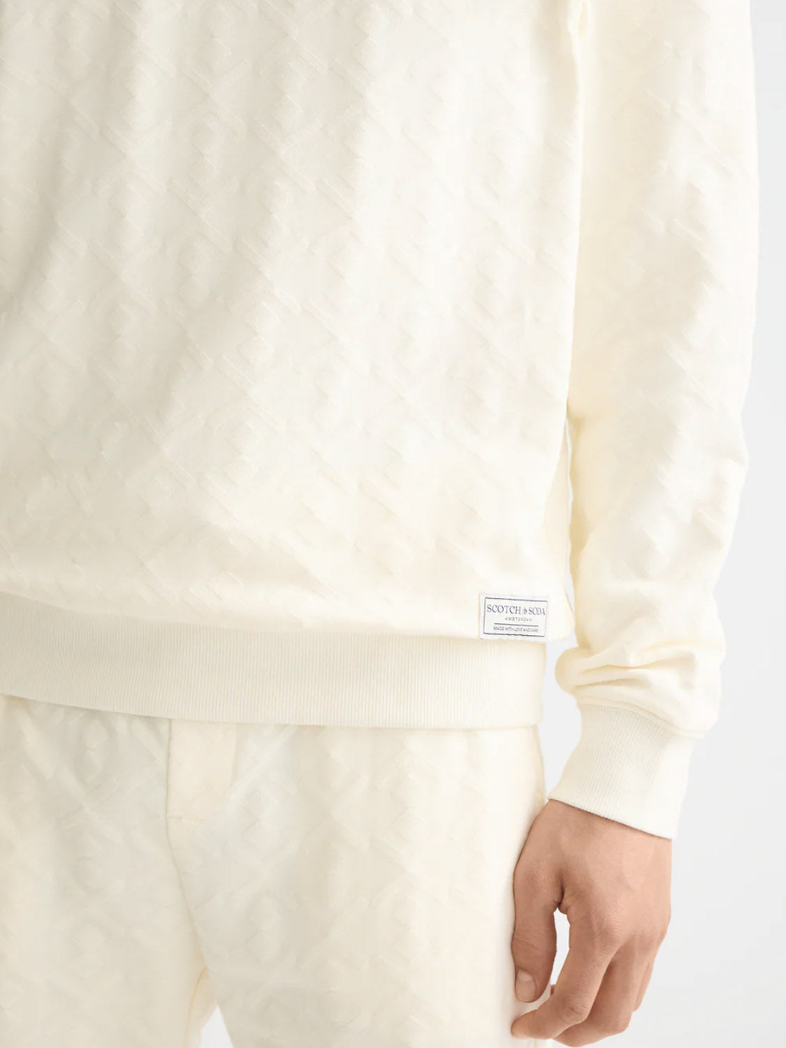 A person wears the Scotch & Soda 181532 Jacquard Terry Sweatshirt in off-white with matching pants, featuring a subtle embossed pattern and a small rectangular label near the sweatshirt's hem.