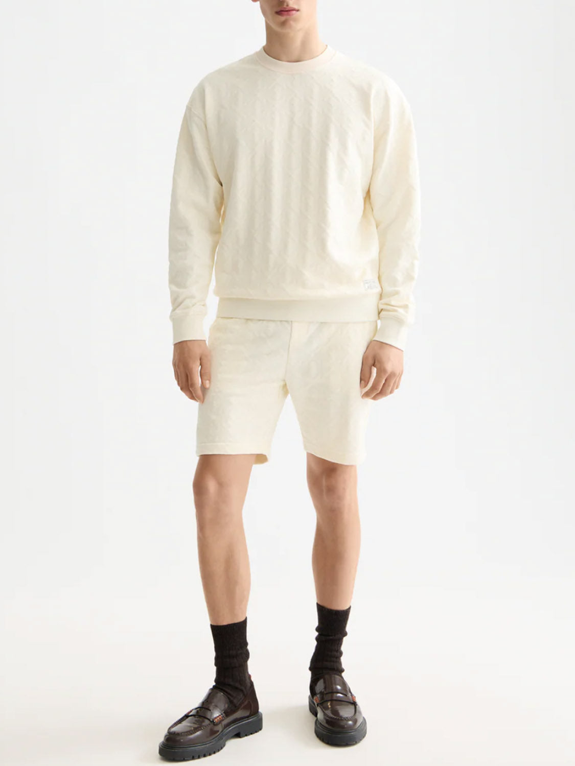 A person stands against a plain background wearing a Scotch & Soda 181532 Jacquard Terry Sweatshirt in off-white with matching shorts, black socks, and dark brown loafers. The person's face is not visible.