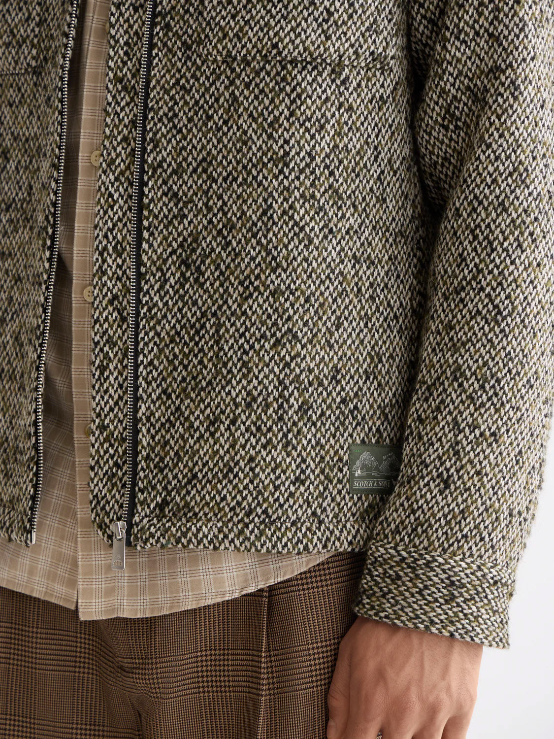 A close-up of a person wearing the Scotch & Soda 181609 Multicolored Tweed Overshirt—a wool-blend jacket with a zipper and small right-side hem label—layered over a beige plaid shirt, paired with brown plaid pants.