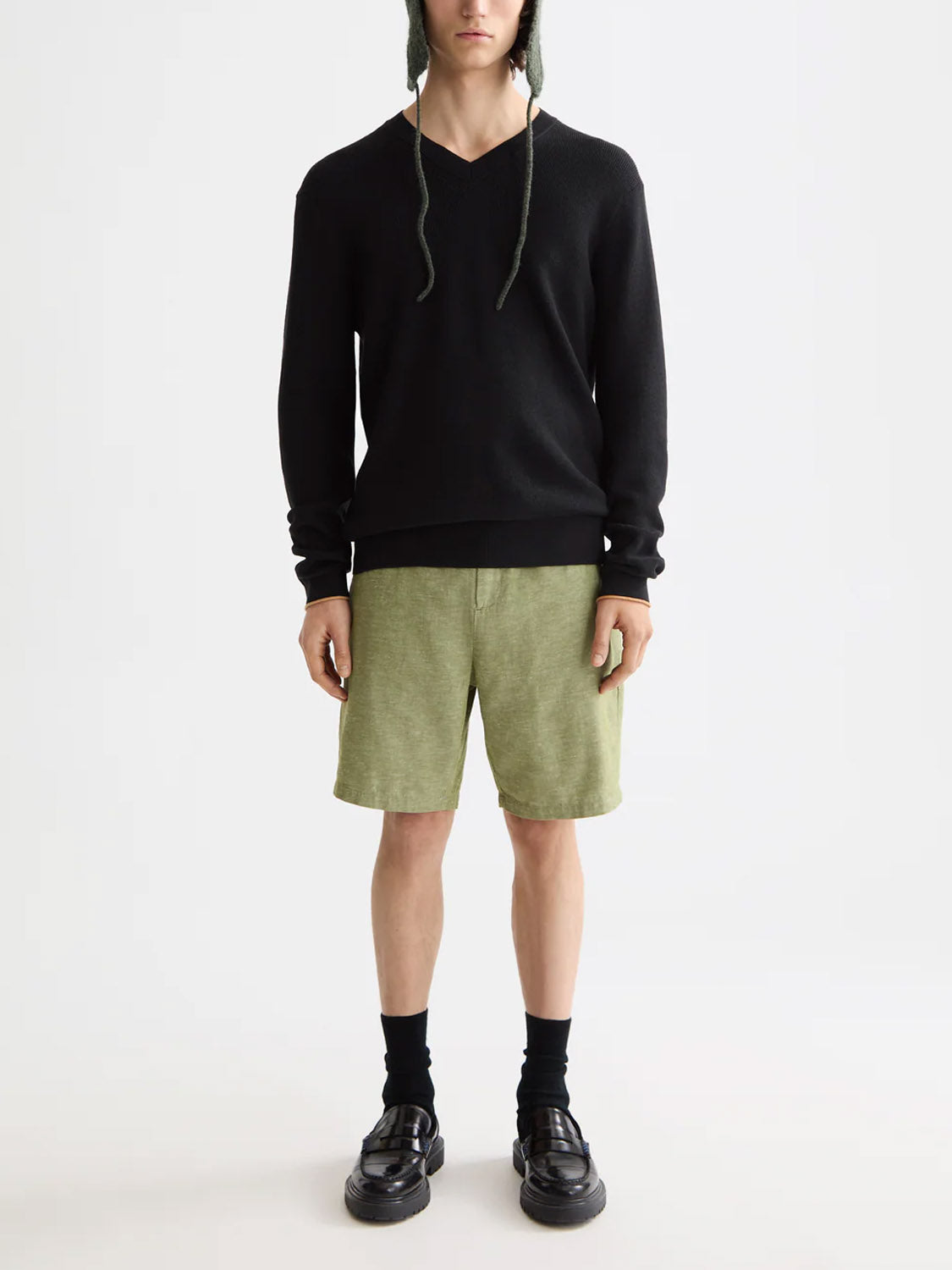 A person stands against a plain background wearing Scotch & Soda's 181773 Cotton Linen V-Neck Pullover, mid-length green textured shorts with an adjustable waistband, black socks and shoes, and a green knit hat with ear flaps and strings.