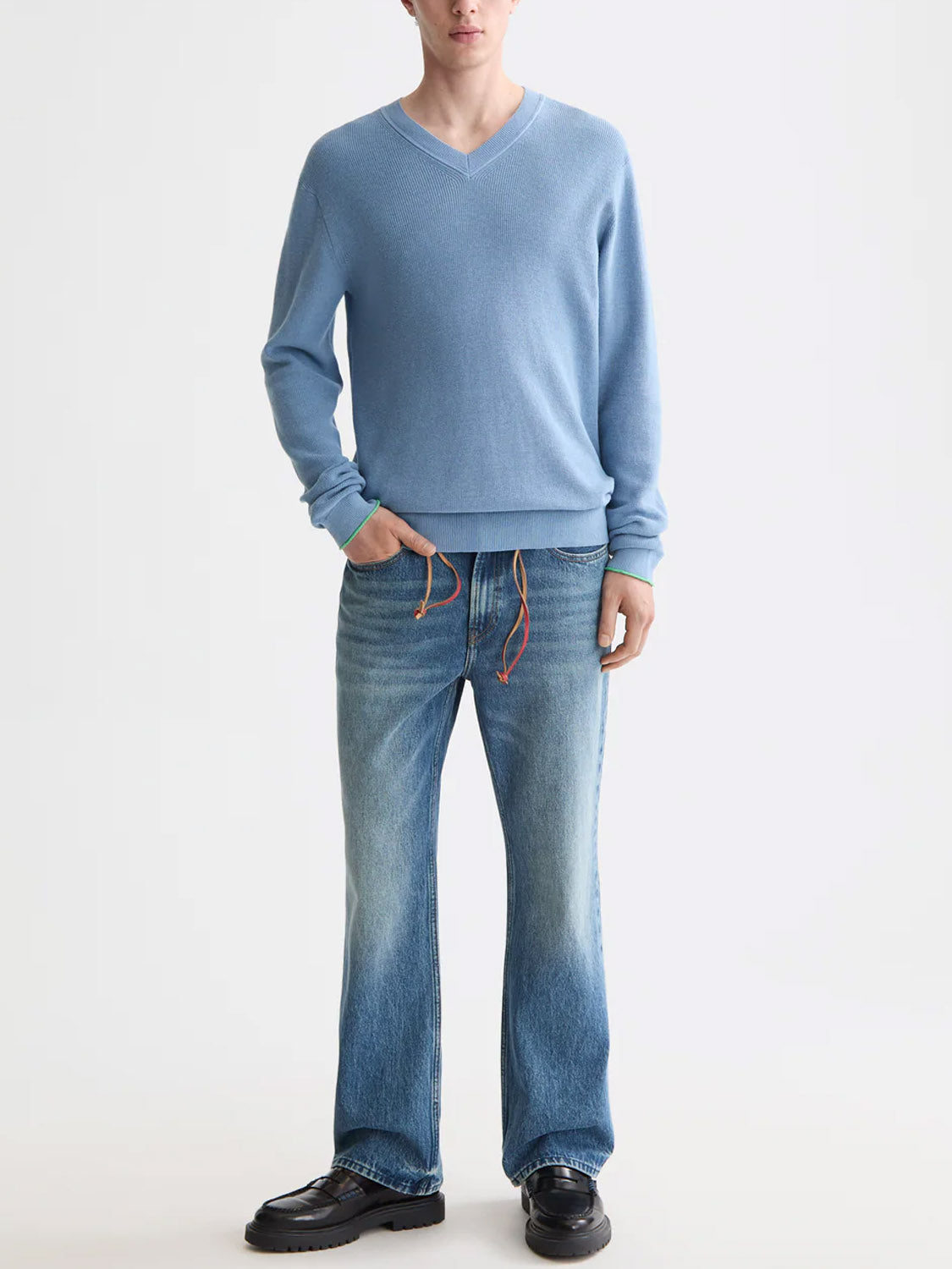 A person wears the Scotch & Soda 181773 Cotton Linen V-Neck Pullover in light blue with faded blue jeans and black shoes, standing against a white background with hands in pockets; their face is partially out of frame.