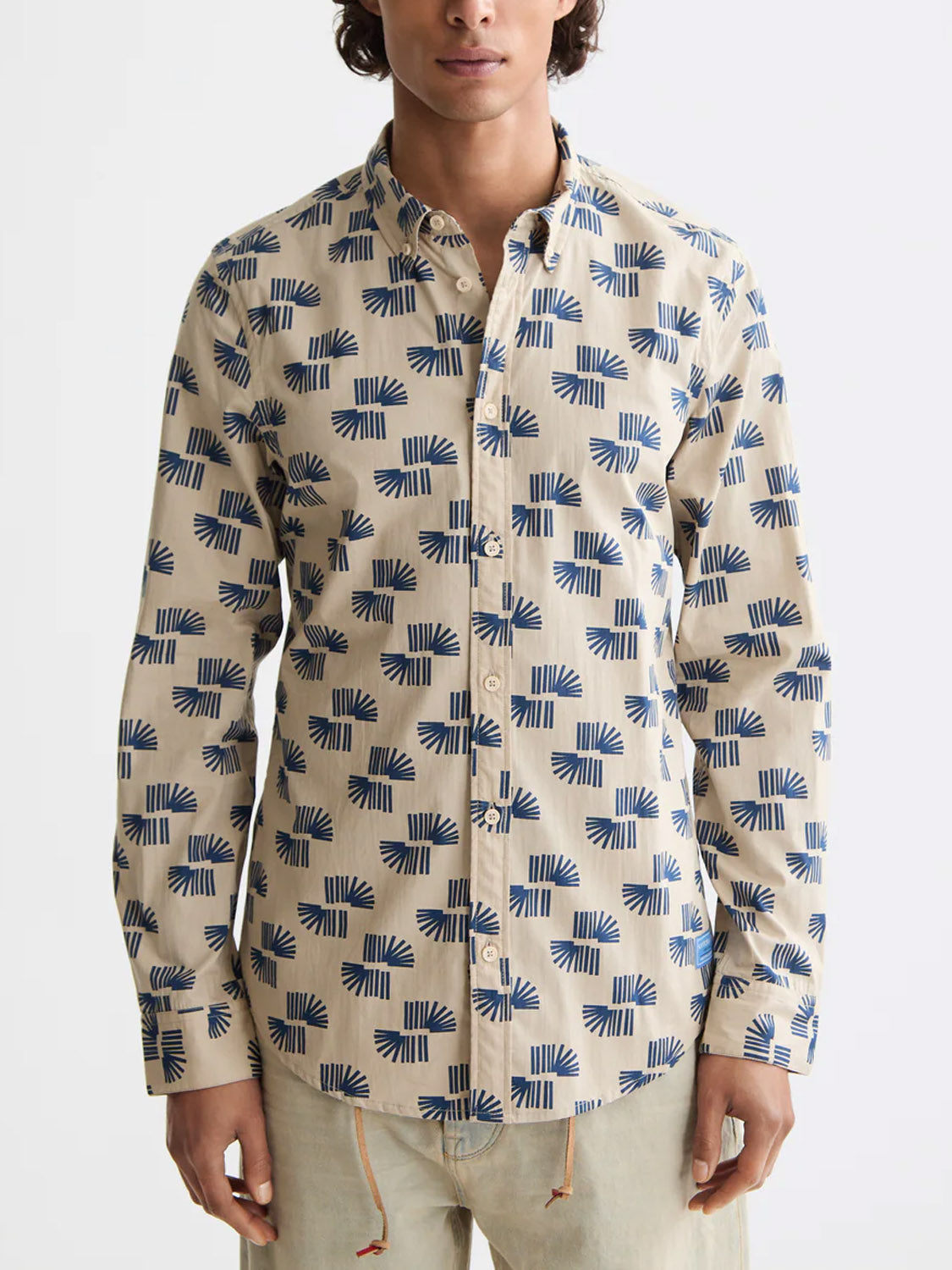 A person wears the Scotch & Soda 181804 Slim Fit Micro All-Over Printed Shirt in beige with a blue geometric square pattern, featuring a collar and long sleeves, made from 100% cotton, against a white background.