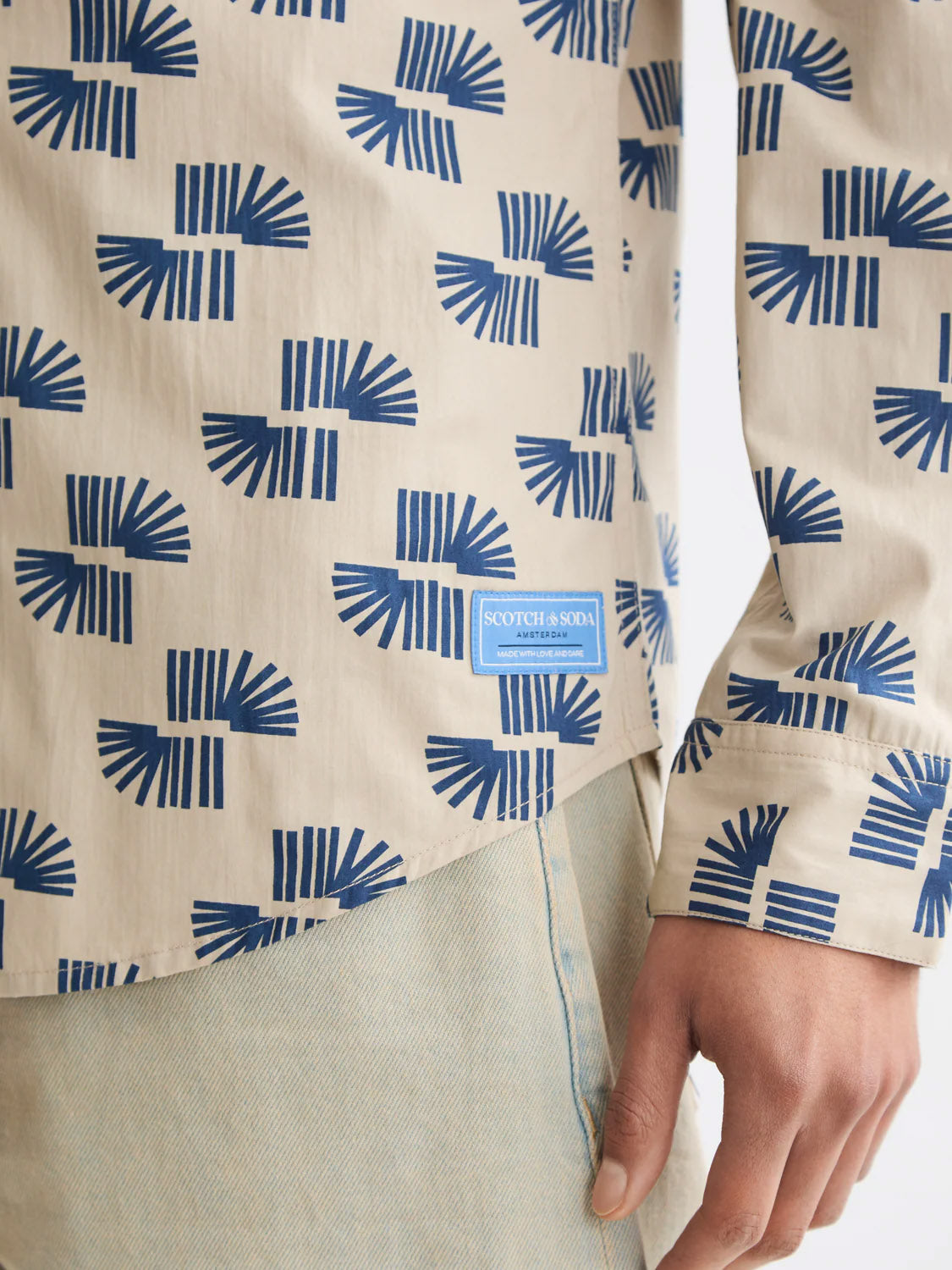 A person wears the Scotch & Soda 181804 Slim Fit Micro All-Over Printed Shirt in beige with a blue geometric pattern and light blue jeans. The Scotch & Soda label is visible, and their left arm hangs at their side.