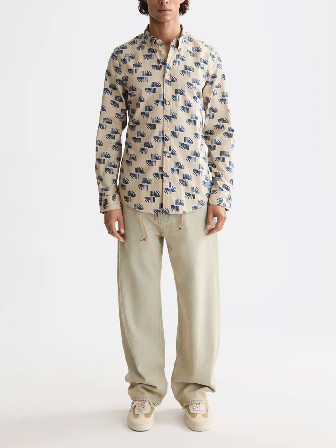 A person faces forward against a plain white background, wearing the Scotch & Soda 181804 Slim Fit Micro All-Over Printed Shirt in beige with blue print, loose light beige pants, and beige sneakers.