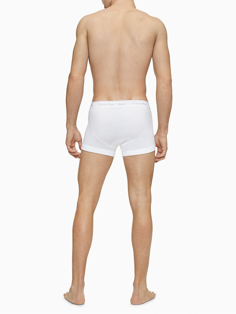 A man stands barefoot facing away from the camera, wearing only Calvin Klein NB4002 3-Pack Cotton Classic Fit Trunks. He is shirtless, and the waistband of the men's underwear features a subtle Calvin Klein logo. The background is plain white.
